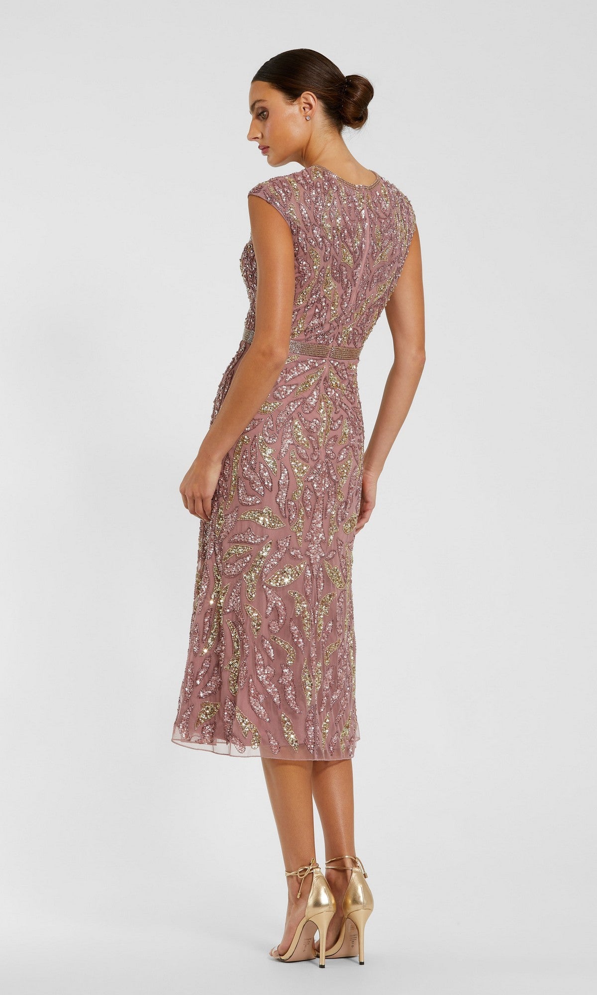 A woman stands in profile with her hair in a low bun, wearing gold high heel sandals and the Mac Duggal 23049 Modest Midi Party Dress—a mauve sheath style featuring intricate beaded embellishments, a fitted waist, and elegant cap sleeves.
